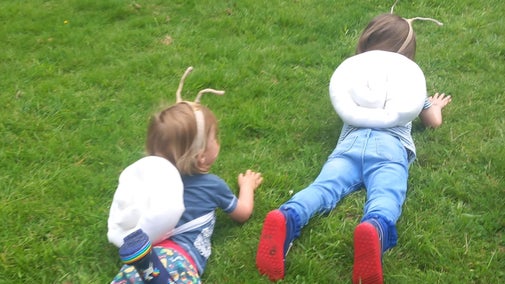 Two children with "snail shells" strapped to their backs and antennae crawling on their bellies on the grass pretending to be snails
